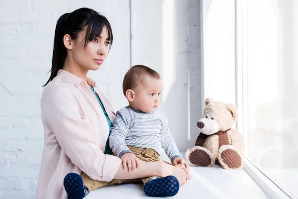 Young mother embracing her little child while he sitting on windowsill and looking through window — Stock Photo