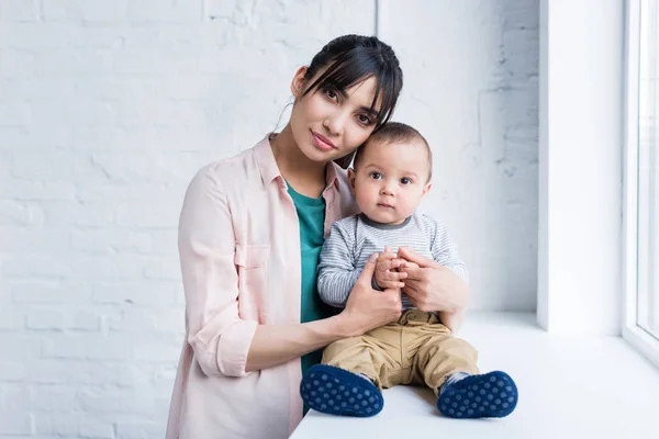 Junge schöne Mutter umarmt ihr kleines Kind, während er auf der Fensterbank sitzt — Stockfoto