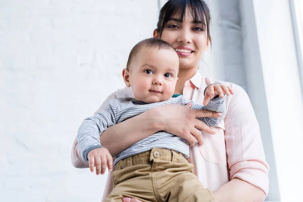 Blick von unten auf eine junge lächelnde Mutter, die ihr kleines Kind trägt und in die Kamera schaut — Stockfoto