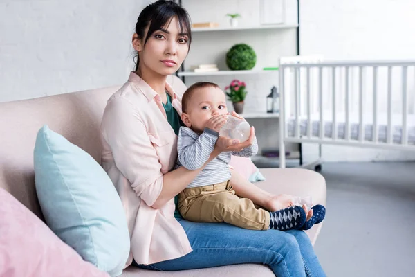 Young beautiful mother feeding her little child with bottle at home and looking at camera — Stock Photo