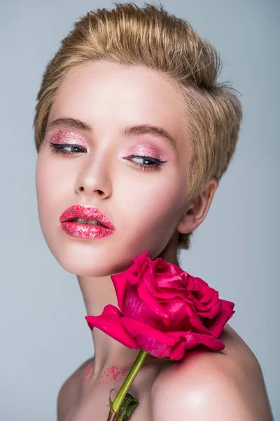 Hermosa mujer con brillo en los labios y rosa roja en el hombro mirando hacia otro lado aislado en gris - foto de stock