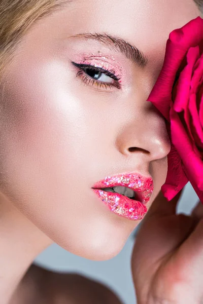 Attractive woman with glitter on lips touching face with red rose — Stock Photo