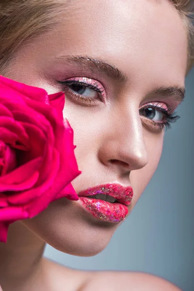 Atractiva mujer con brillo en los labios sosteniendo rosa roja y mirando a la cámara aislada en gris - foto de stock