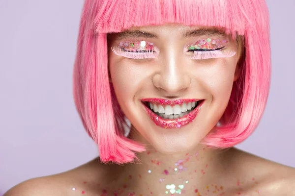 Mujer atractiva sonriente con pelo rosa, maquillaje brillante y pestañas largas aisladas en violeta - foto de stock