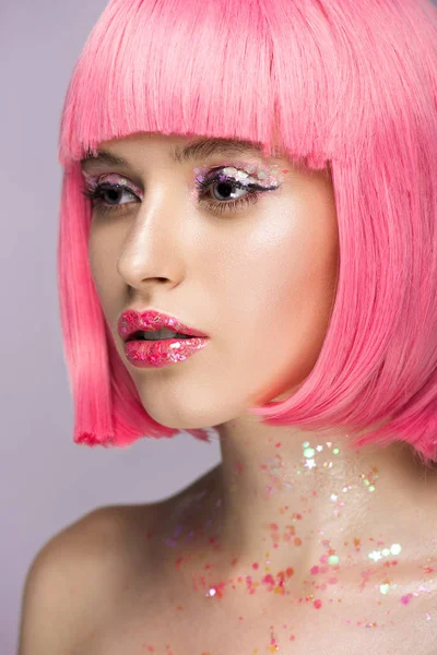 Atractiva mujer con pelo rosa y maquillaje con brillo mirando hacia otro lado aislado en violeta - foto de stock