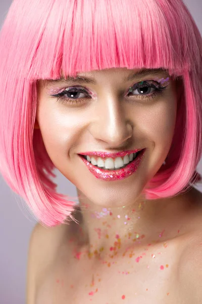 Mujer atractiva sonriente con pelo rosa y maquillaje con brillo mirando a la cámara aislada en violeta - foto de stock