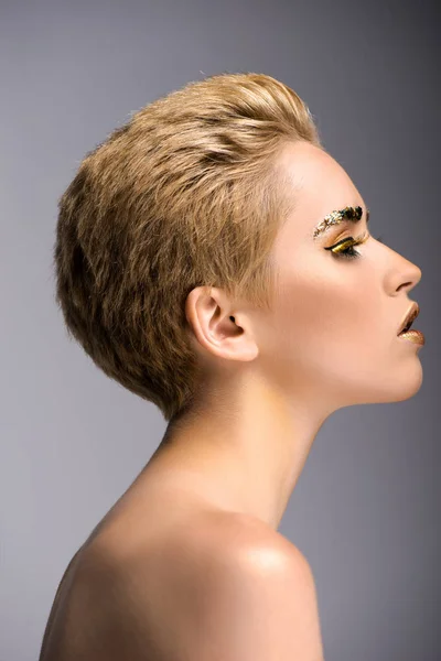 Vue latérale d'une jolie femme aux paillettes dorées sur le visage et aux cheveux courts isolés sur le gris — Photo de stock