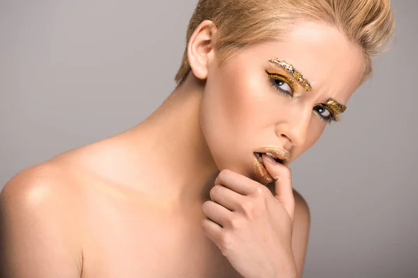 Attractive woman with golden glitter on face biting thumb isolated on grey — Stock Photo