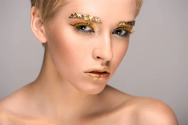 Hermosa mujer con maquillaje brillante dorado mirando a la cámara aislada en gris - foto de stock