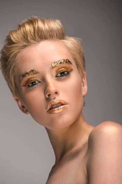 Vue à angle bas d'une jolie femme avec des paillettes dorées sur le visage regardant la caméra isolée sur le gris — Photo de stock