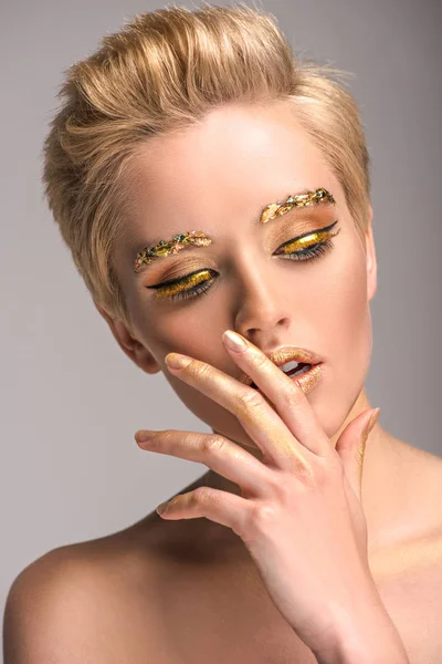 Atractiva mujer con brillo dorado en la cara tocando los labios aislados en gris - foto de stock