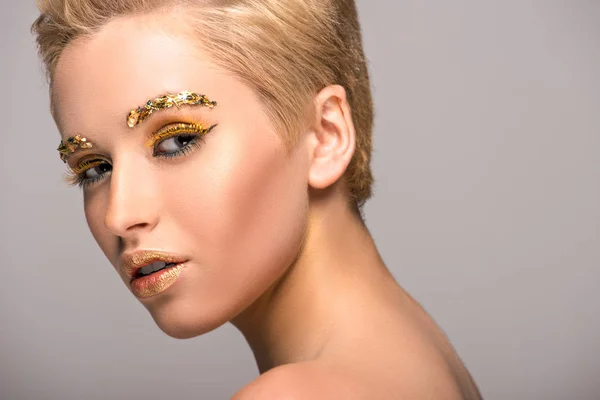 Jolie femme avec des paillettes dorées sur le visage en regardant la caméra isolée sur gris — Photo de stock