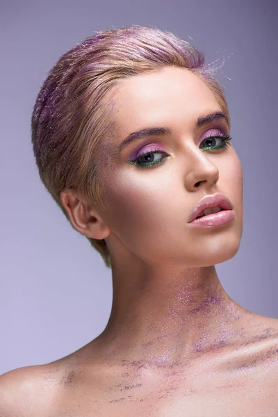 Приваблива жінка з фіолетовим блиском на шиї і коротким волоссям дивиться на камеру ізольовано на фіолетовому — стокове фото