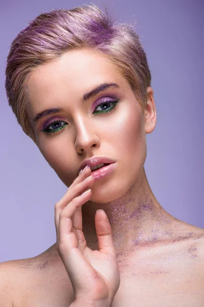 Jolie femme avec des paillettes violettes sur le cou et les cheveux courts touchant les lèvres isolées sur violet — Photo de stock