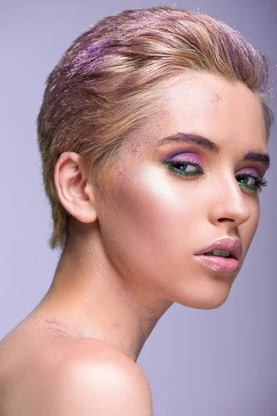 Atractiva mujer con brillo violeta en el cuello y pelo corto mirando a la cámara aislada en violeta - foto de stock