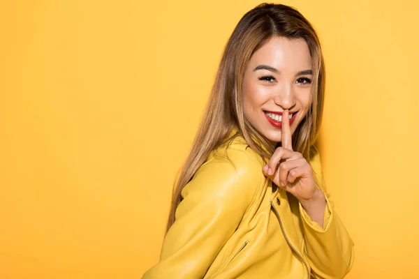 Sorridente asiatica modello femminile chiusura bocca per dito isolato su sfondo giallo — Foto stock