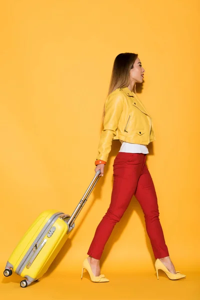 Side view of stylish asian female traveler carrying wheeled bag on yellow background — Stock Photo