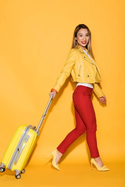 Viajero femenino sonriente llevando un bolso de ruedas sobre fondo amarillo - foto de stock