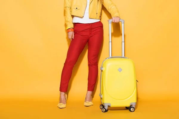 Обрезанный снимок стильной женщины-путешественницы, стоящей с колёсной сумкой на желтом фоне — стоковое фото
