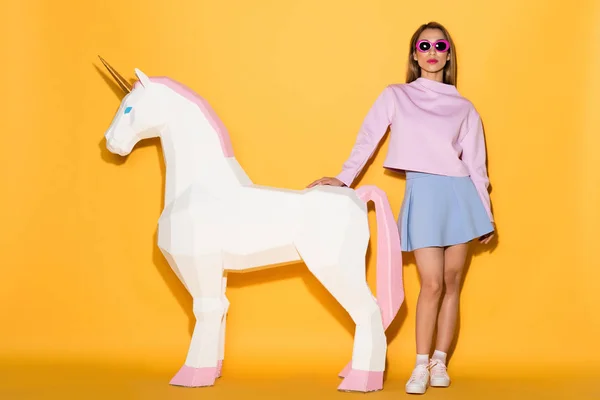 Élégant modèle féminin asiatique en lunettes de soleil et licorne décorative sur fond jaune — Photo de stock