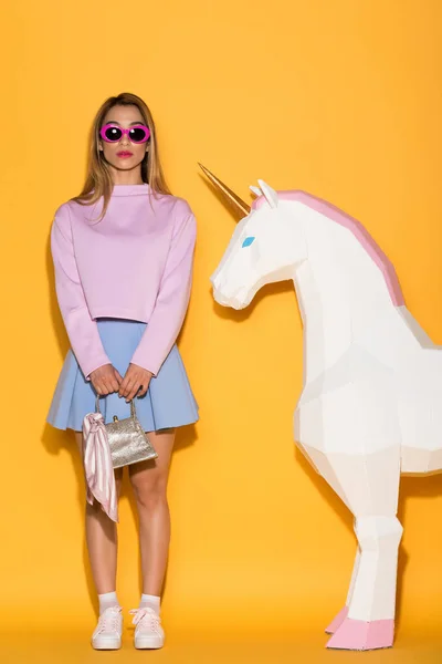 Elegante asiático feminino modelo em óculos de sol segurando bolsa e unicórnio decorativo no fundo amarelo — Fotografia de Stock