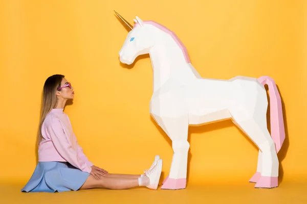Side view of asian female model in sunglasses sitting on floor and decorative unicorn on yellow background — Stock Photo