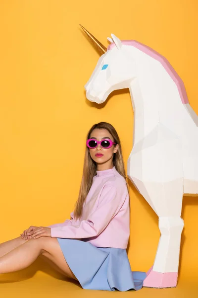 Modelo femenino asiático con estilo en gafas de sol y unicornio decorativo sobre fondo amarillo - foto de stock