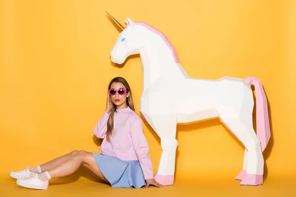 Decorative unicorn and stylish asian female model in sunglasses sitting on floor on yellow background — Stock Photo