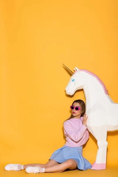 Asian female model in sunglasses sitting on floor and decorative unicorn on yellow background — Stock Photo