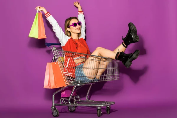 Beautiful happy young woman in sunglasses holding paper bags and sitting in shopping trolley on violet — Stock Photo