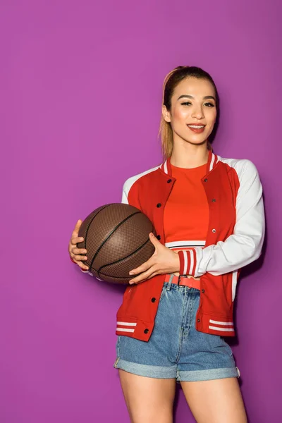 Schöne asiatische Mädchen hält Basketballball und lächelt in die Kamera isoliert auf violett — Stockfoto