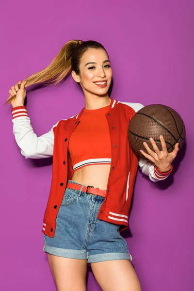 Красива весела азіатська дівчина тримає баскетбол і посміхається на камеру ізольована на фіолетовому — стокове фото