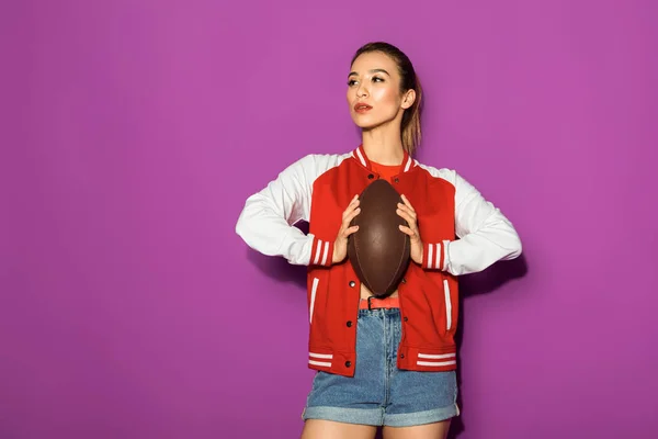 Beautiful asian woman holding rugby ball and looking away isolated on violet — Stock Photo