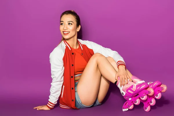 Красивая улыбающаяся азиатская девушка в роликовых коньках сидит и смотрит в сторону изолированы на фиолетовый — стоковое фото