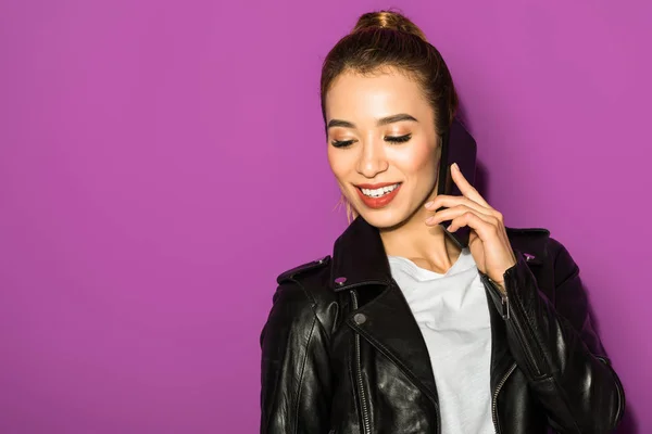 Beautiful smiling asian girl talking by smartphone and looking down isolated on violet — Stock Photo