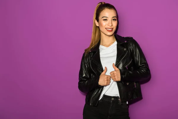 Beautiful stylish asian girl in leather jacket smiling at camera isolated on violet — Stock Photo