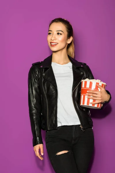 Bonito sorrindo asiático menina no couro jaqueta segurando pipoca e olhando para longe isolado no violeta — Fotografia de Stock