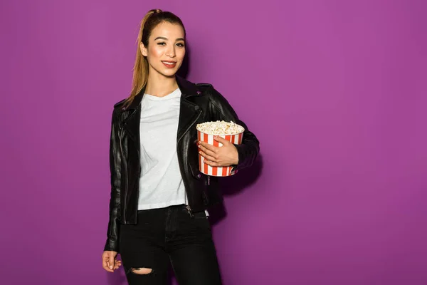Beautiful stylish asian girl holding box of popcorn and smiling at camera isolated on violet — Stock Photo
