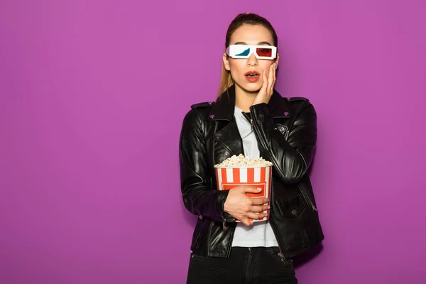 Шокированная молодая женщина в 3D очки едят попкорн и глядя на камеру изолированы на фиолетовый — стоковое фото