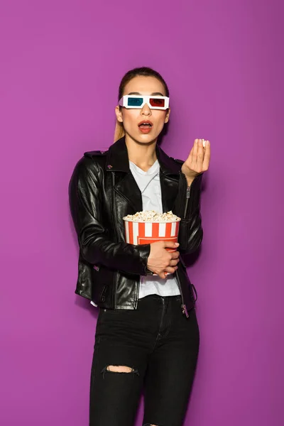 Shocked asian girl in leather jacket and 3d glasses eating popcorn and looking at camera isolated on violet — Stock Photo