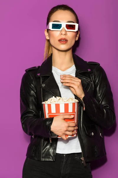 Beautiful asian girl in leather jacket and 3d glasses eating popcorn and looking at camera isolated on violet — Stock Photo