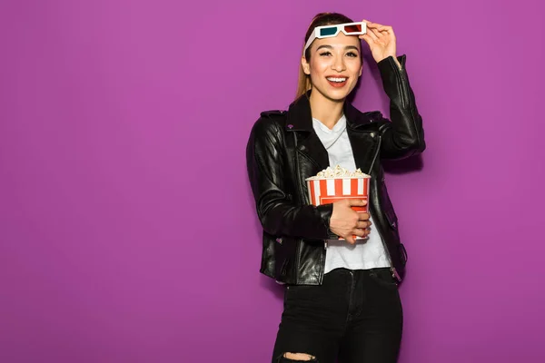 Bella felice asiatico ragazza in 3d bicchieri holding popcorn e sorridente a fotocamera isolato su viola — Foto stock