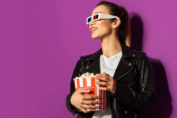 Красива усміхнена азіатська дівчина в 3d окулярах тримає попкорн і дивиться ізольовано на фіолетовий — стокове фото