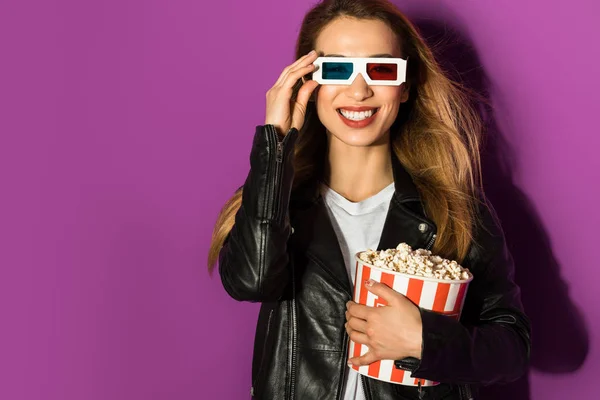 Красива молода жінка в 3d окулярах тримає коробку попкорну і посміхається на камеру ізольовано на фіолетовому — стокове фото