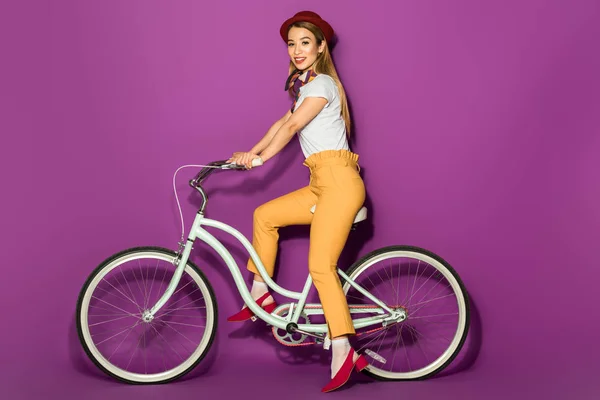 Красива стильна азіатська дівчина їде на велосипеді і посміхається на камеру ізольовано на фіолетовому — стокове фото