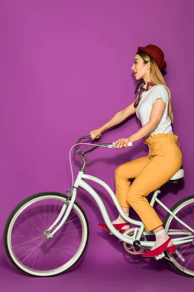 Вид збоку приваблива стильна азіатська дівчина їде на велосипеді і дивиться ізольовано на фіолетовому — стокове фото