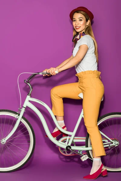 Вид в полный рост счастливой стильной девушки, катающейся на велосипеде и улыбающейся камере на фиолетовом фоне — стоковое фото
