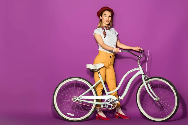 Hermosa elegante chica asiática de pie con bicicleta y sonriendo a la cámara aislado en violeta - foto de stock