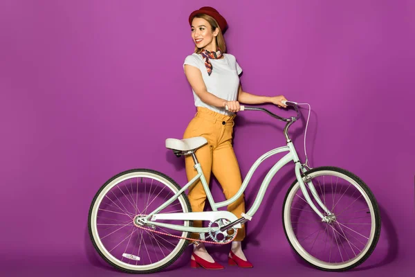 Вид в полный рост красивой улыбающейся стильной девушки, стоящей с велосипедом и отводящей вдаль взгляд на фиолетовый — стоковое фото