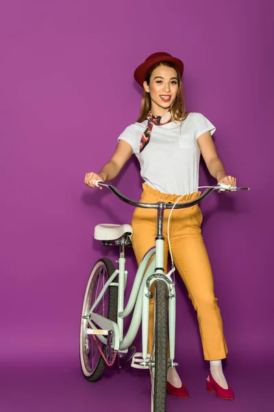 Full length view of beautiful stylish asian woman standing with bicycle and soring at câmera isolated on violet — Fotografia de Stock